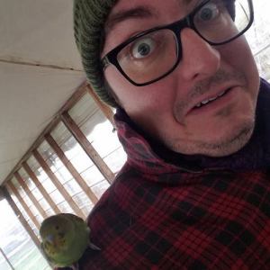 A selfie of Ben, a nonbinary white person wearing a red plaid flannel jacket and a green beanie with a bird on their shoulder.