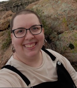 A cropped selfie of Andi, an androgynous white person with a buzzed haircut, glasses, a white shirt and black overalls.