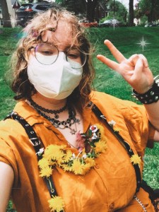 A selfie of Ly, a trans white person wearing a yellow top with flowers accessorized. He is wearing a studded bracelet and a face mask.